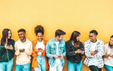 A posed photo of a diverse group of young people, laughing and talking together. Yellow backdrop wall.